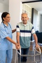 Smiling multiracial female physiotherapist assisting senior man in walking with walker at home Royalty Free Stock Photo