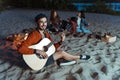 Smiling musician playing guitar while his friends sitting behind on sandy beach