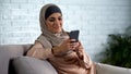 Smiling muslim woman using smartphone at home, chatting in social media