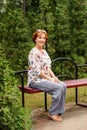 Smiling aged lady sits on a bench in the greenery parks in linen clothes