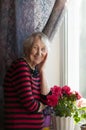 Smiling old woman near the window. Happy senior woman sitting near window with flowers.