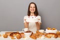 Smiling optimistic woman wearing white T-shirt isolated over gray background sitting at festive table among homemade desserts
