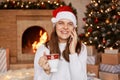 Smiling optimistic young adult woman wearing white sweater and santa claus hat, posing near fireplace and xmas tree in festive Royalty Free Stock Photo