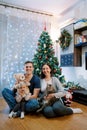 Smiling parents with a little girl and a cat on their knees sit near the Christmas tree on the floor Royalty Free Stock Photo