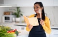 Smiling pensive young black lady cook in apron with wooden spoon, reads recipe book, thinks about new dish Royalty Free Stock Photo