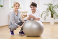 Smiling physiotherapist with elderly woman laying on exercising ball during physical therapy