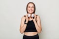 Smiling positive cheerful slim woman with headphones wearing sportswear posing against gray background lookign at camera with