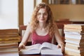 Smiling positive girl sitting at table in library, having several bunch of books around, looking directly at camera, touching Royalty Free Stock Photo