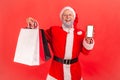 Smiling positive gray bearded santa claus holding and showing paper bags and empty screen smartphone, shopping online