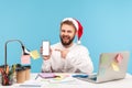 Smiling positive man office worker in santa claus hat pointing at smartphone with empty display sitting at workplace with laptop, Royalty Free Stock Photo