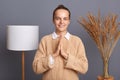 Smiling positive woman wearing beige sweater posing in stylish room concentrating her mind, keeping hands namaste gesture,