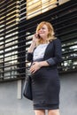 A smiling pregnant businesswoman standing in front of a modern building having a phone call