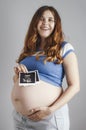 Smiling pregnant cute young caucasian woman standing against grey studio background and holding an ultrasound black and white scan Royalty Free Stock Photo