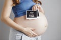 Smiling pregnant cute young caucasian woman standing against grey studio background and holding an ultrasound black and white scan Royalty Free Stock Photo