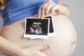 Smiling pregnant cute young caucasian woman standing against grey studio background and holding an ultrasound black and white scan Royalty Free Stock Photo