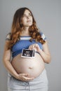 Smiling pregnant cute young caucasian woman standing against grey studio background and holding an ultrasound black and white scan Royalty Free Stock Photo