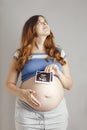 Smiling pregnant cute young caucasian woman standing against grey studio background and holding an ultrasound black and white scan Royalty Free Stock Photo