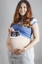 Smiling pregnant cute young caucasian woman standing against grey studio background and holding an ultrasound black and white scan Royalty Free Stock Photo