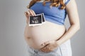 Smiling pregnant cute young caucasian woman standing against grey studio background and holding an ultrasound black and white scan Royalty Free Stock Photo