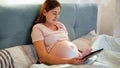 Smiling pregnant woman in pajamas using tablet computer and typing message while lying in bed at night Royalty Free Stock Photo