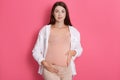 Smiling pregnant woman wearing white shirt and leggins, looking at camera and touches her belly, expectant mother standing Royalty Free Stock Photo