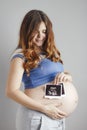 Smiling pregnant young woman with red hair and blue eyes on grey studio background and holding an ultrasound black and white scan Royalty Free Stock Photo