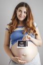 Smiling pregnant young woman with red hair and blue eyes on grey studio background and holding an ultrasound black and white scan Royalty Free Stock Photo