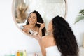 Smiling pretty arab millennial curly lady in towel applying blush on face, looking in mirror, enjoy daily procedure
