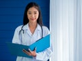 Smiling pretty Asian woman doctor portrait standing on blue wood and white curtain background in hospital room. Royalty Free Stock Photo