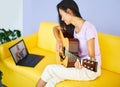 Smiling pretty female learning to play guitar watches a video lesson online using laptop