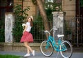 Pretty female in red dress holds flowers with blue vintage bike near her standing in front of beautiful old house Royalty Free Stock Photo