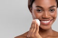 Smiling pretty millennial african american female wiping her face with cotton pad, isolated on gray background, close up