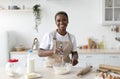 Smiling pretty millennial african american woman chef in apron making dough in light minimalist kitchen interior