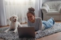 Smiling pretty mixed-race woman chilling on carpet floor with dog using laptop Royalty Free Stock Photo