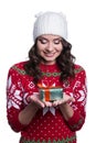 Smiling pretty young woman wearing colorful knitted sweater with christmas ornament and hat, holding christmas gift. Royalty Free Stock Photo