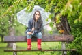 Smiling pretty woman holding umbrella on her shoulder sits on a bench back-rest in the park in the rain