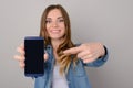 Smiling pretty woman showing black empty screen of her smartphone and pointing on it with her finger; isolated on grey background Royalty Free Stock Photo