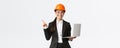 Smiling professional asian female engineer or architect at construction, wearing safety helmet and suit, pointing finger Royalty Free Stock Photo