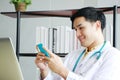 Smiling Professional Asian Psychiatrist or neurologist doctor man is relaxing with smartphone at computer desk in clinical office Royalty Free Stock Photo