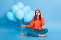 Smiling red haired young girl wearing casual sweater and jeans, holding bunch of blue helium balloons, having excellent mood,