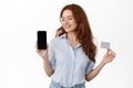 Smiling redhead businesswoman showing empty phone screen and credit card. Office lady looking satisfied at smartphone Royalty Free Stock Photo