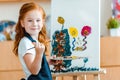 smiling redhead child standing near painting on canvas in art school.