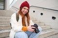 Smiling redhead girl photographer, checks her shots, holds camera and looks at screen, takes photos outdoors, walks