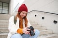 Smiling redhead girl photographer, checks her shots, holds camera and looks at screen, takes photos outdoors, walks