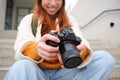 Smiling redhead girl photographer, checks her shots, holds camera and looks at screen, takes photos outdoors, walks