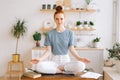 Smiling redhead young woman is meditating while sitting on desk at home office, looking at camera. Royalty Free Stock Photo