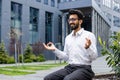 Smiling and relaxed young Indian male businessman sitting on bench in lotus position near offices, meditating and Royalty Free Stock Photo