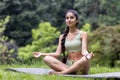 Smiling and relaxed young Indian woman in sportswear sitting in the park on a mat in the lotus position and doing yoga Royalty Free Stock Photo