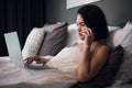 Smiling relaxed young woman using mobile phone and laptop in bed at home Royalty Free Stock Photo
