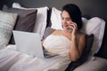 Smiling relaxed young woman using mobile phone and laptop in bed at home Royalty Free Stock Photo